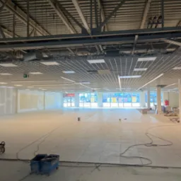 Wnętrze komercyjne w trakcie układania płytek podłogowych, widoczne narzędzia glazurnicze i surowa ściana przygotowana do wykończenia.