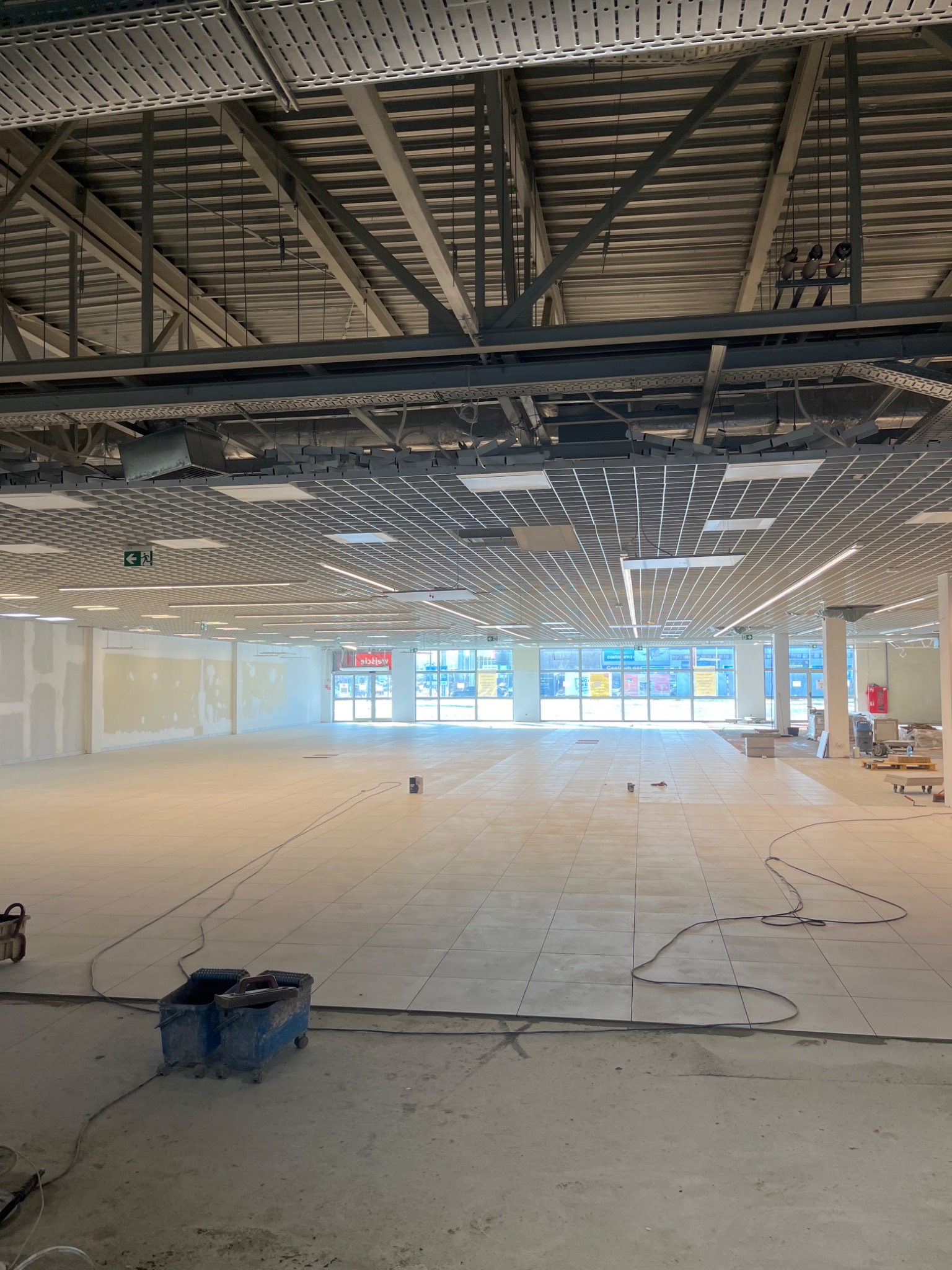 Wnętrze komercyjne w trakcie układania płytek podłogowych, widoczne narzędzia glazurnicze i surowa ściana przygotowana do wykończenia.