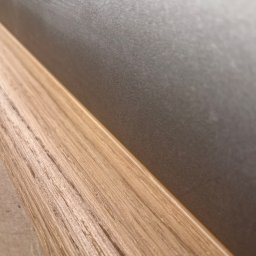 JOBI_BUD - Szczegółowe ujęcie połączenia drewnianej listwy przypodłogowej z szarą ścianą o delikatnej fakturze.