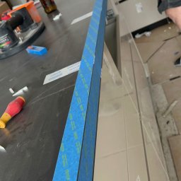 JOBI_BUD - Przygotowanie do fugowania płytek: pionowy pasek niebieskiej taśmy malarskiej zabezpiecza krawędź blatu przed zabrudzeniem, widoczny fragment glazury, narzędzia na blacie i dłoń pracownika w rękawicy.