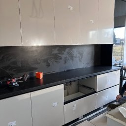 JOBI_BUD - Montaż nowoczesnej zabudowy kuchennej: beżowe szafki z połyskiem, czarny blat imitujący marmur, widoczne narzędzia glazurnicze i pozostałości folii ochronnej na frontach.