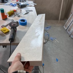 JOBI_BUD - Płytka ceramiczna z wyfrezowanym rowkiem trzymana w dłoni na tle warsztatu glazurnika z narzędziami: szlifierką kątową, gąbką i taśmą malarską.