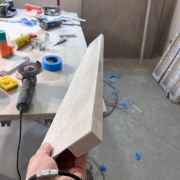 JOBI_BUD - Przygotowana do montażu, długa, beżowa płytka ceramiczna trzymana w dłoni, w tle stół roboczy z narzędziami: szlifierką kątową, gąbką, taśmą malarską i klejem, oraz fragment ściany z ułożonymi...