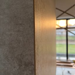 JOBI_BUD - Fragment ściany z nałożoną gładzią o fakturze betonu, widoczne odbicie światła, w tle okno z widokiem na zieleń i rusztowanie.