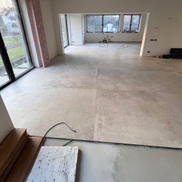 JOBI_BUD - Wnętrze w trakcie prac wykończeniowych: ułożona jasna, wielkoformatowa podłoga z płytek imitujących kamień, widoczne niezabezpieczone kable, przygotowane panele podłogowe, oraz fragment ocieplenia...