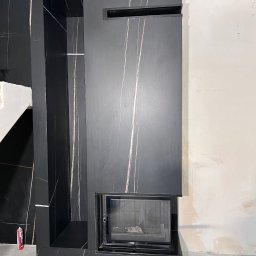 JOBI_BUD - Częściowo wykończony czarny kominek z marmurowym wzorem, widoczne niedoskonałości na ścianie i podłodze, wskazujące na trwający remont.