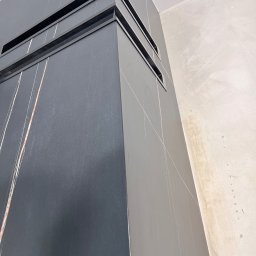 JOBI_BUD - Narożnik obudowany ciemnoszarą płytką imitującą kamień z jasnymi żyłkami, widoczne wnęki wentylacyjne, surowa ściana w tle.
