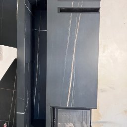 JOBI_BUD - Nowoczesna obudowa kominka z ciemnego marmuru z jasnymi żyłami, z wnęką na drewno i minimalistycznymi wstawkami, w trakcie prac wykończeniowych.