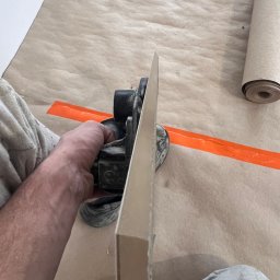 JOBI_BUD - Ręka trzymająca szlifierkę do gładzi na tle zabezpieczonej podłogi z rolką papieru i pomarańczową taśmą maskującą.