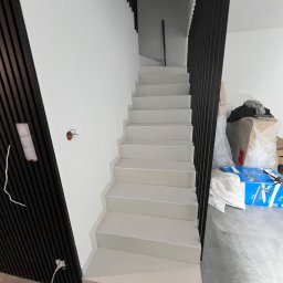 JOBI_BUD - Schody wewnętrzne z ciemną balustradą i panelami ściennymi, świeżo pomalowane na biało, z widocznymi puszkami elektrycznymi i kablami, oraz kartonami w tle.