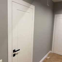 JOBI_BUD - Białe drzwi z czarną klamką w minimalistycznym wnętrzu z szarą ścianą i drewnopodobną podłogą.