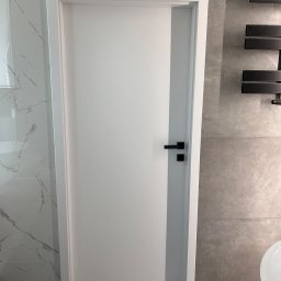 JOBI_BUD - Białe drzwi z czarną klamką w łazience wykończonej płytkami imitującymi marmur i beton, widoczny czarny grzejnik drabinkowy.