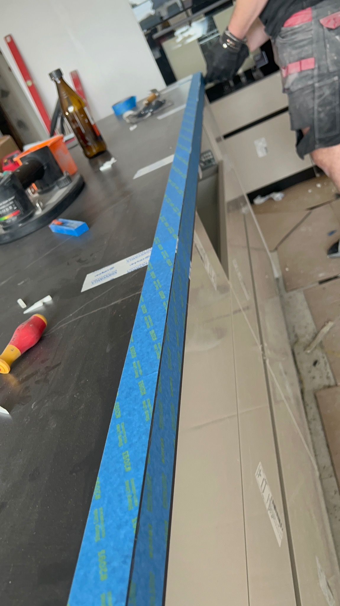 Przygotowanie do fugowania płytek: pionowy pasek niebieskiej taśmy malarskiej zabezpiecza krawędź blatu przed zabrudzeniem, widoczny fragment glazury, narzędzia na blacie i dłoń pracownika w rękawicy.