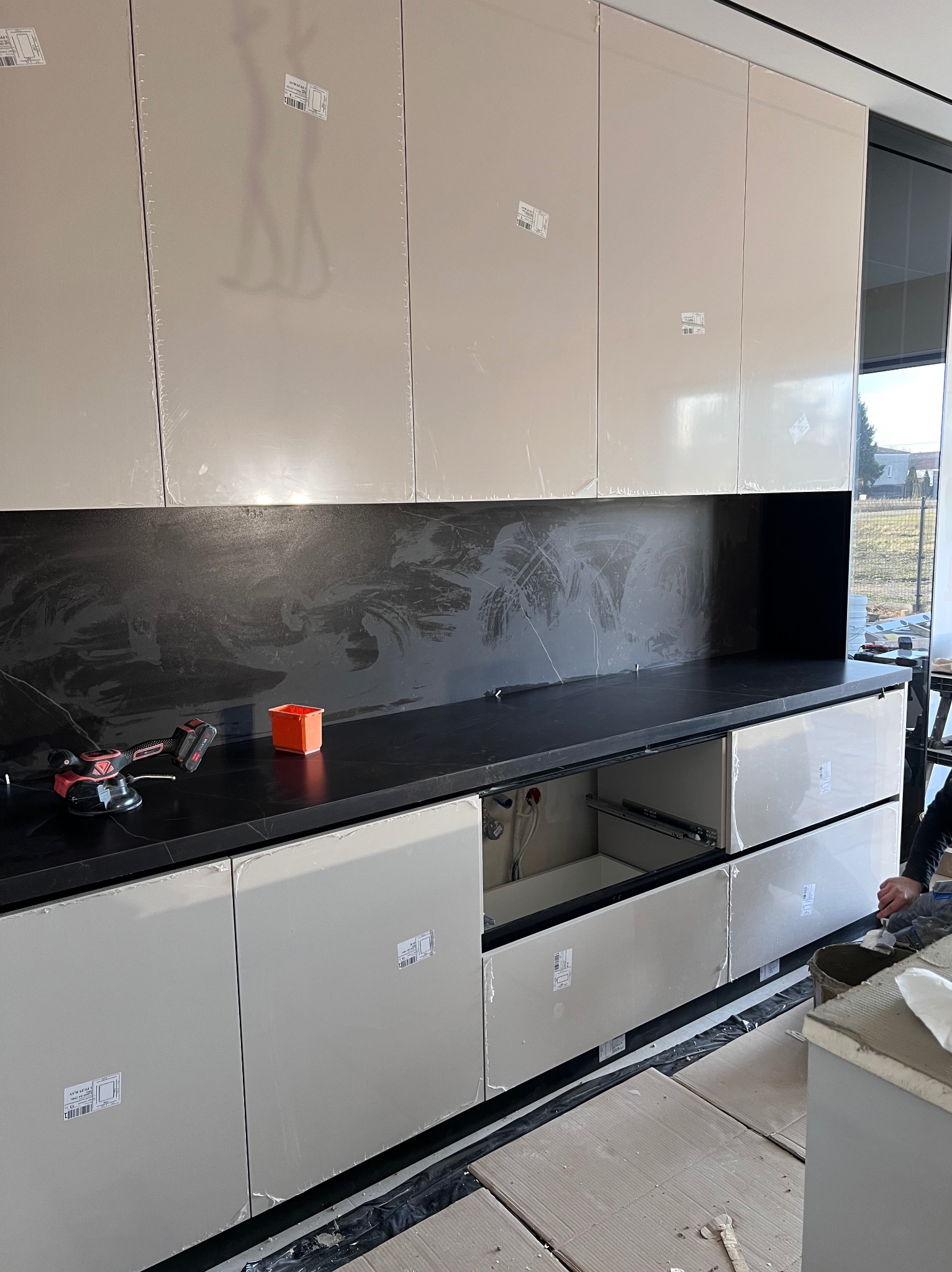 Montaż nowoczesnej zabudowy kuchennej: beżowe szafki z połyskiem, czarny blat imitujący marmur, widoczne narzędzia glazurnicze i pozostałości folii ochronnej na frontach.