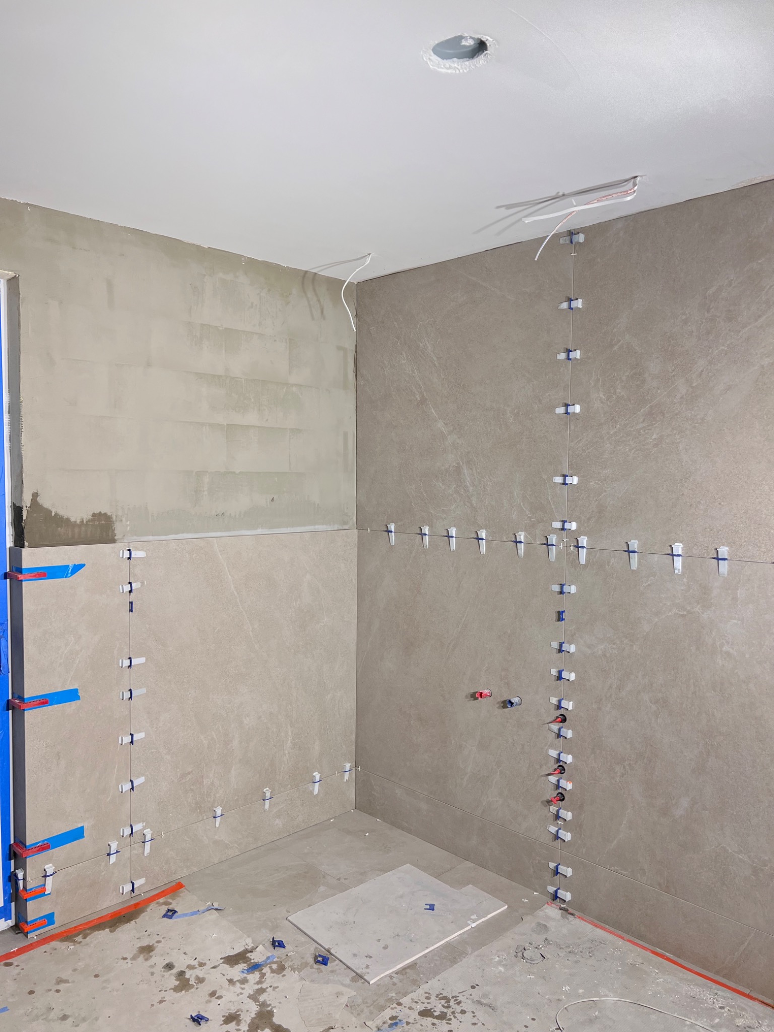 Wnętrze łazienki w trakcie remontu, widoczne częściowo położone płytki imitujące kamień na ścianach i podłodze, użyte klipsy poziomujące, fragment ściany z nałożonym klejem, wystające przewody...