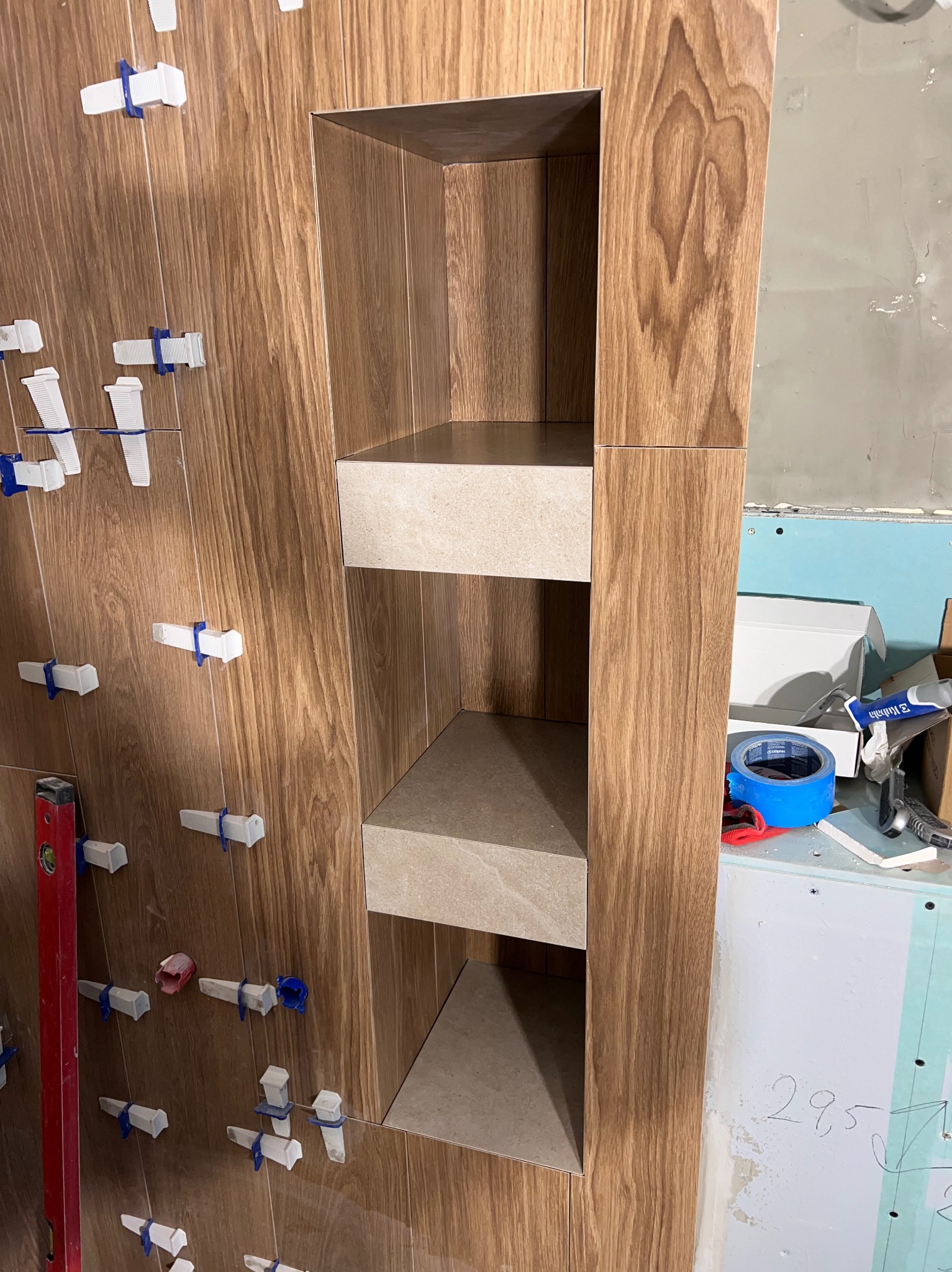 Płytki imitujące drewno na ścianie z wnęką, widoczne poziomica, niebieska taśma malarska i białe klipsy dystansowe do płytek podczas montażu.