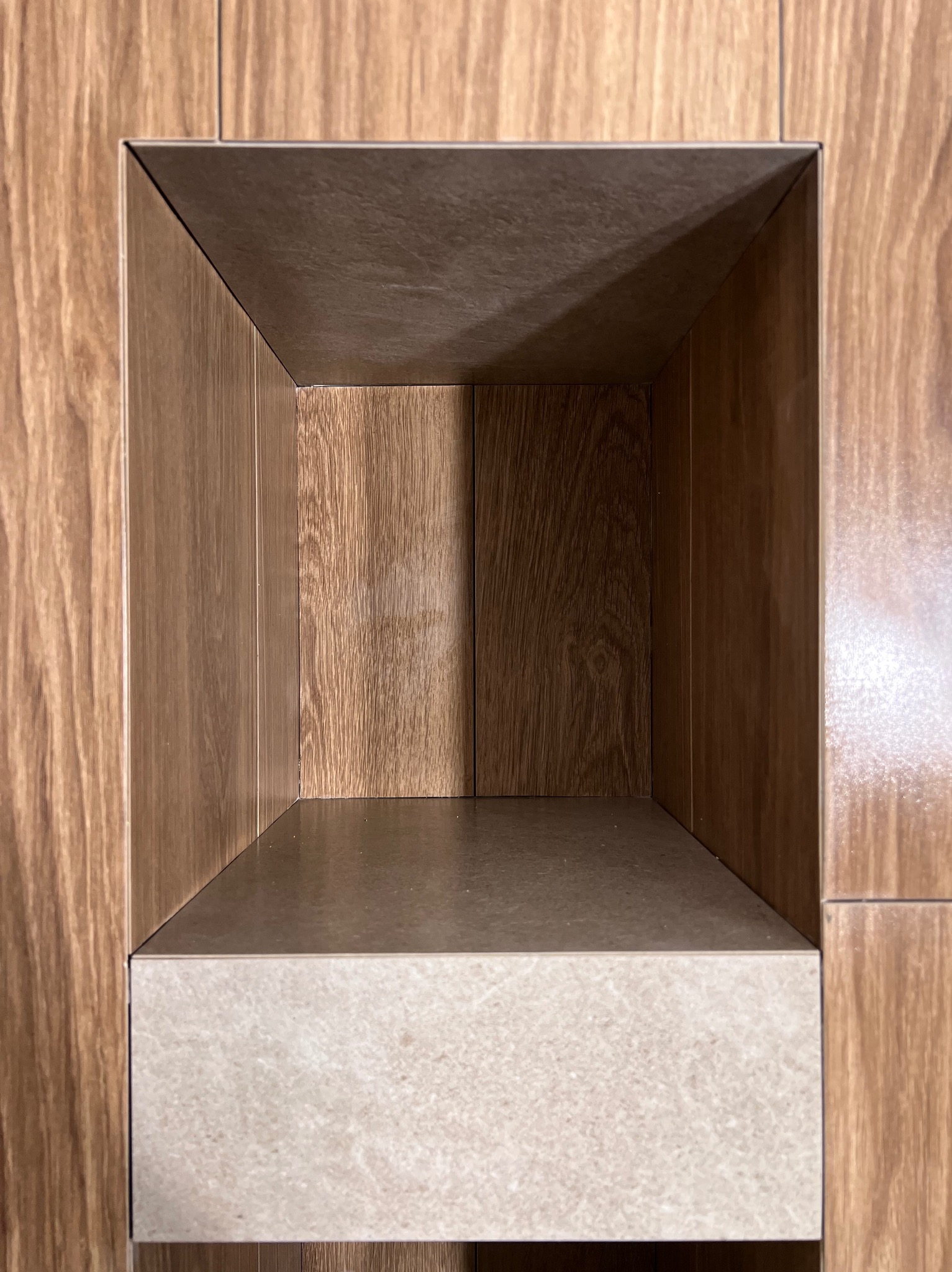 Wnęka w ścianie wyłożona płytkami imitującymi drewno, z półką z jasnego kamienia, tworząca minimalistyczny element dekoracyjny w łazience.