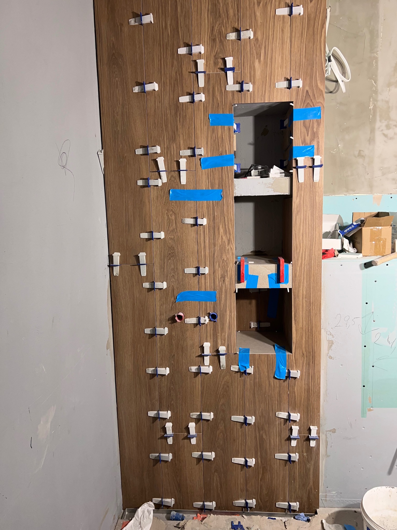 Montaż pionowych, drewnopodobnych płytek z użyciem klipsów poziomujących i niebieskiej taśmy, widoczne wnęki w ścianie, surowy stan wykończenia pomieszczenia.