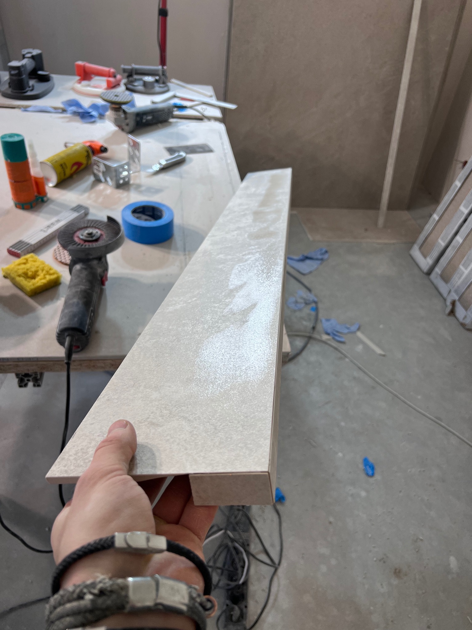 Płytka ceramiczna z wyfrezowanym rowkiem trzymana w dłoni na tle warsztatu glazurnika z narzędziami: szlifierką kątową, gąbką i taśmą malarską.