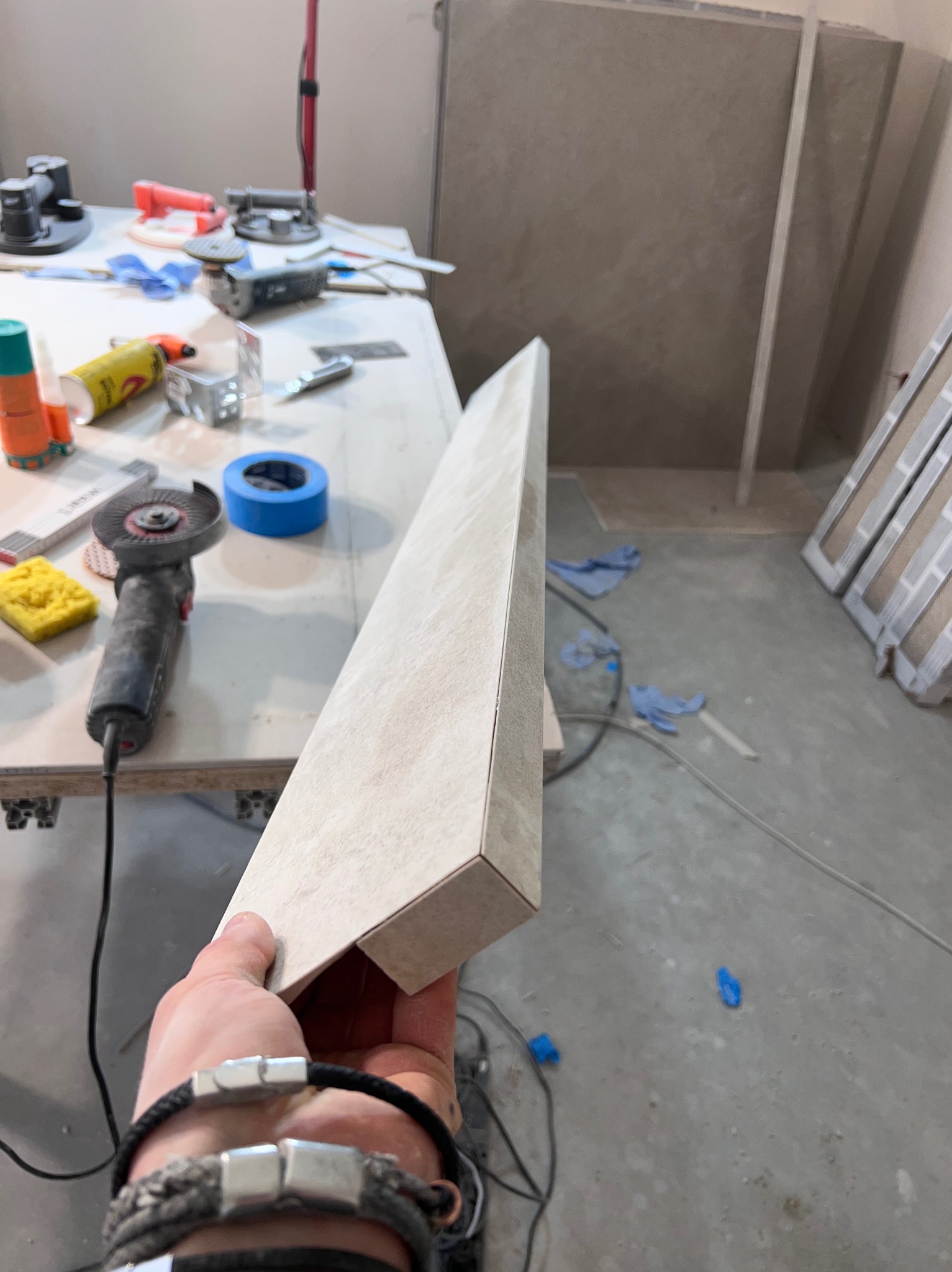 Przygotowana do montażu, długa, beżowa płytka ceramiczna trzymana w dłoni, w tle stół roboczy z narzędziami: szlifierką kątową, gąbką, taśmą malarską i klejem, oraz fragment ściany z ułożonymi...