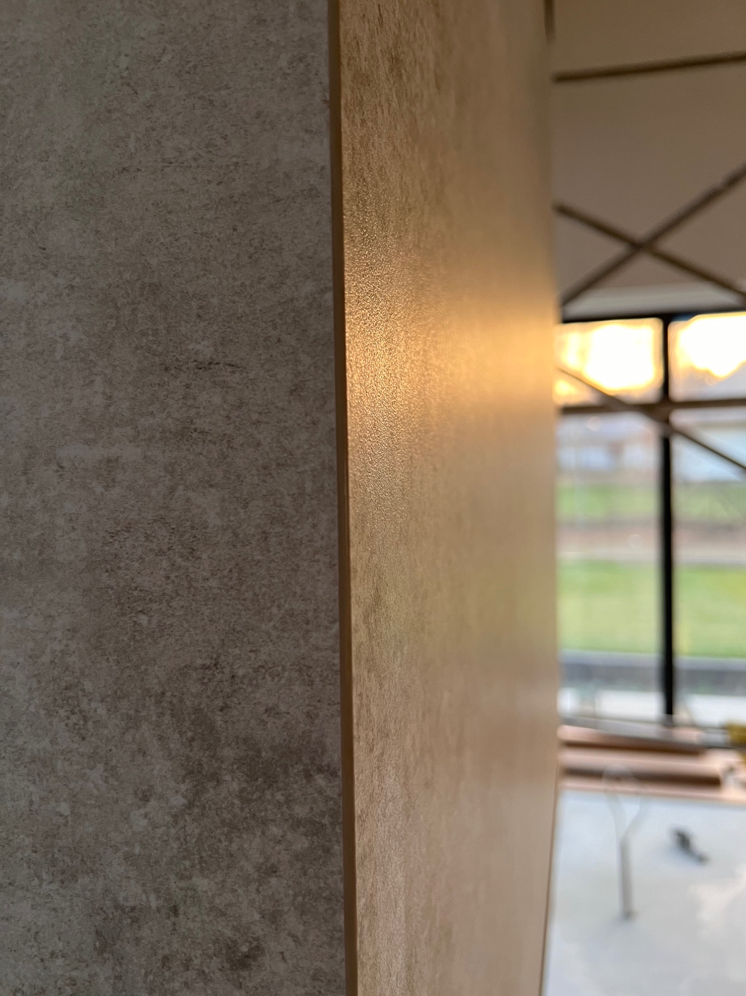 Fragment ściany z nałożoną gładzią o fakturze betonu, widoczne odbicie światła, w tle okno z widokiem na zieleń i rusztowanie.