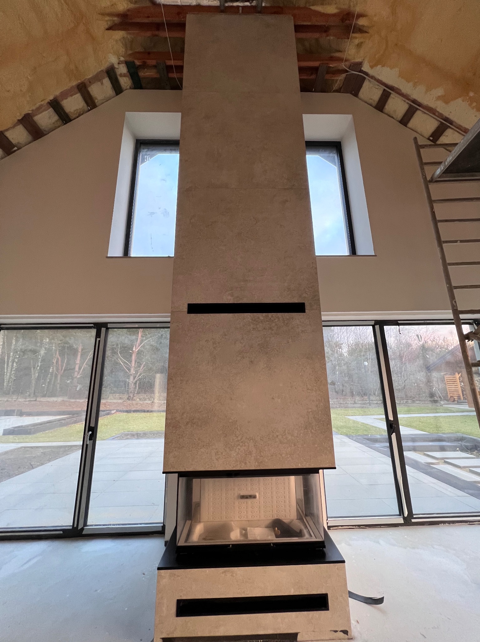 Wnętrze w trakcie wykończenia: kominek obłożony płytami w odcieniach beżu, widoczne okna i surowe wykończenie poddasza z drewnianą konstrukcją.