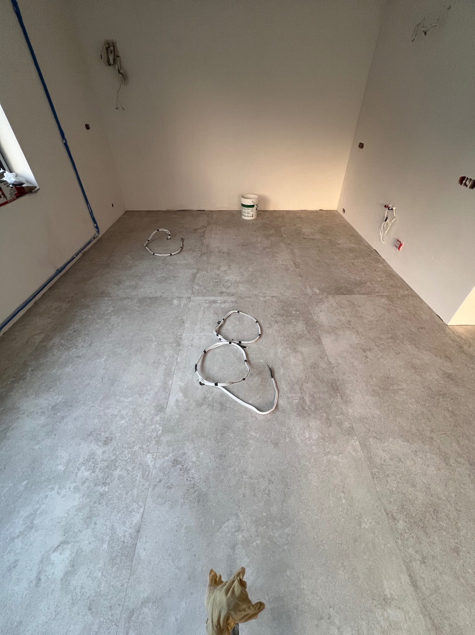 Szare płytki podłogowe w trakcie montażu, widoczne kable grzewcze ułożone na podłodze, ściany przygotowane do malowania, puszka farby w rogu pomieszczenia.