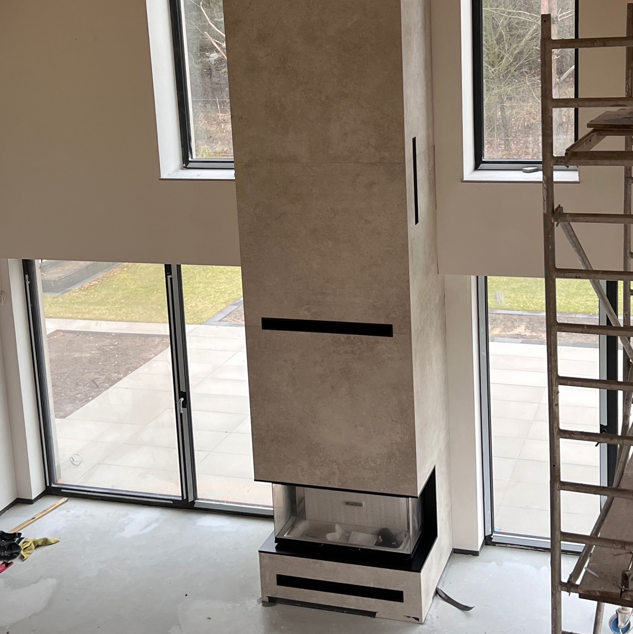 Wysoki, obłożony płytami kominek z czarnymi akcentami, widoczny w surowym wnętrzu z dużymi oknami i rusztowaniem, podłoga w trakcie prac wykończeniowych.