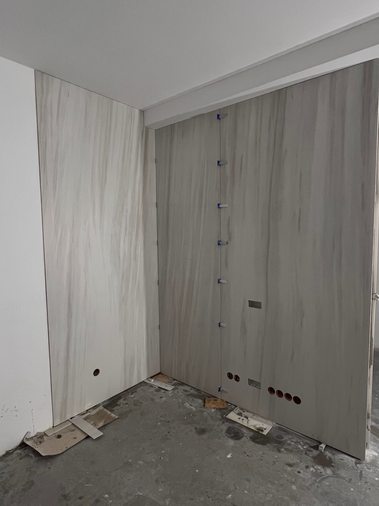 Świeżo położone, pionowe płytki imitujące marmur na ścianie łazienki, z widocznymi niebieskimi krzyżykami dystansowymi i otworami na instalacje, surowy beton na podłodze.