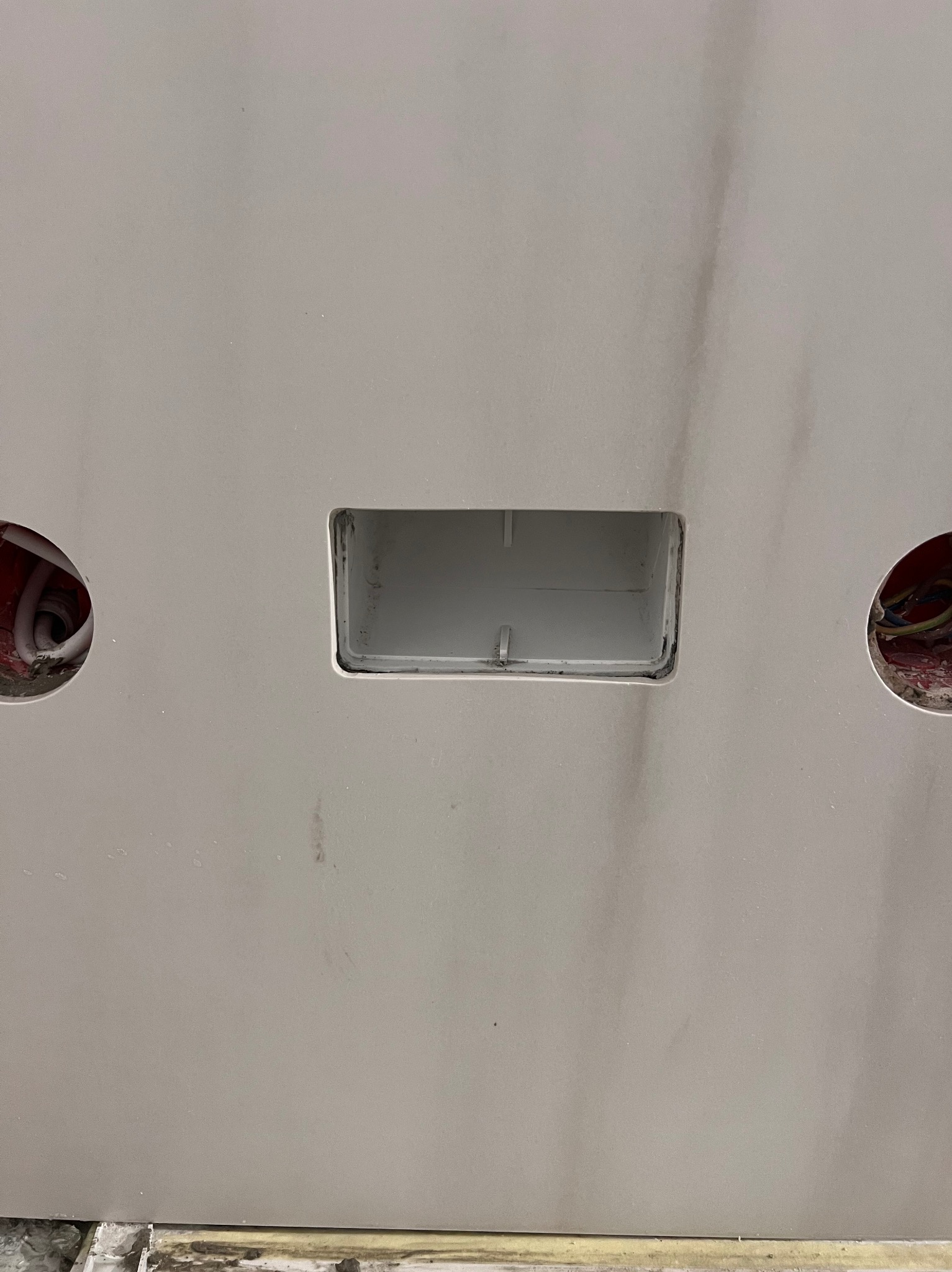 Szara ściana z dwoma okrągłymi otworami z widocznymi przewodami i jednym prostokątnym otworem na puszkę elektryczną.