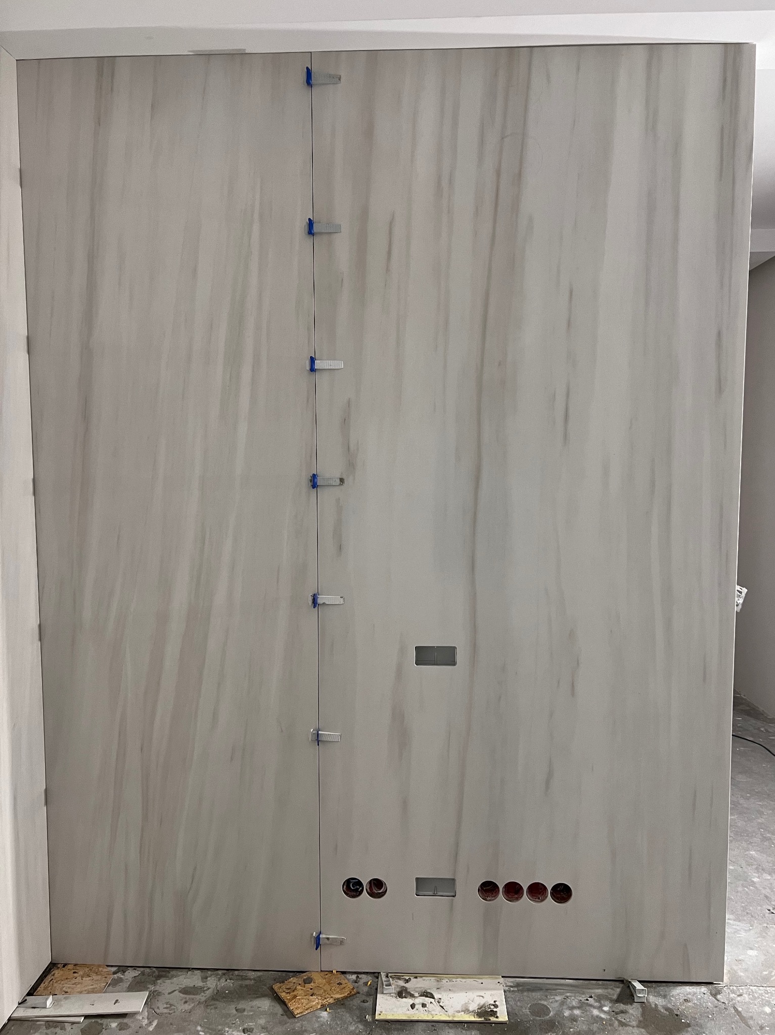 Układanie płytek imitujących marmur na ścianie, widoczne otwory na gniazdka i niebieskie klipsy poziomujące, podłoga zabezpieczona kartonem.