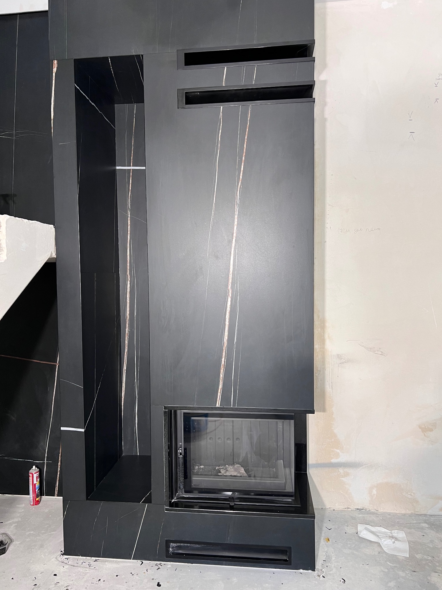 Częściowo wykończony czarny kominek z marmurowym wzorem, widoczne niedoskonałości na ścianie i podłodze, wskazujące na trwający remont.