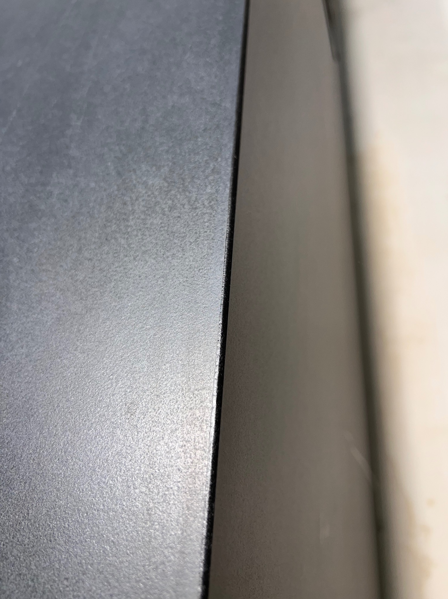 Fragment szarej, matowej powierzchni z widoczną krawędzią i delikatną fakturą, oświetlonej z boku, z jasnym tłem w prawym górnym rogu.