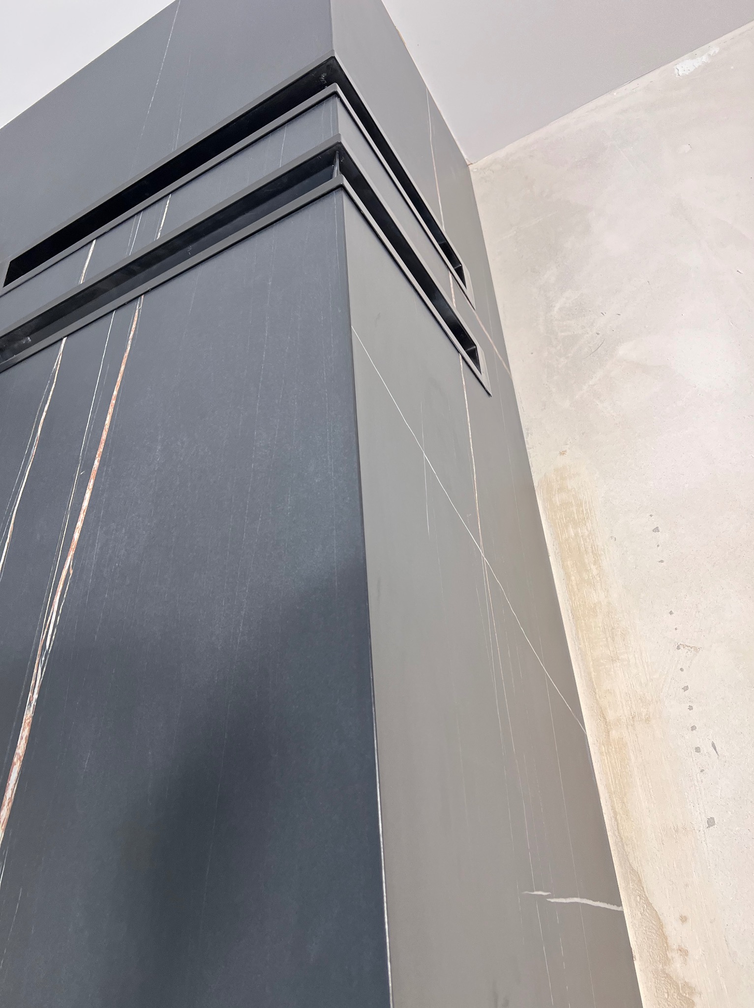 Narożnik obudowany ciemnoszarą płytką imitującą kamień z jasnymi żyłkami, widoczne wnęki wentylacyjne, surowa ściana w tle.