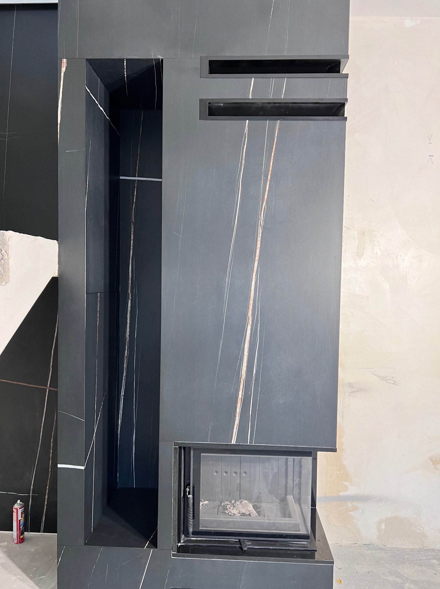 Nowoczesna obudowa kominka z ciemnego marmuru z jasnymi żyłami, z wnęką na drewno i minimalistycznymi wstawkami, w trakcie prac wykończeniowych.