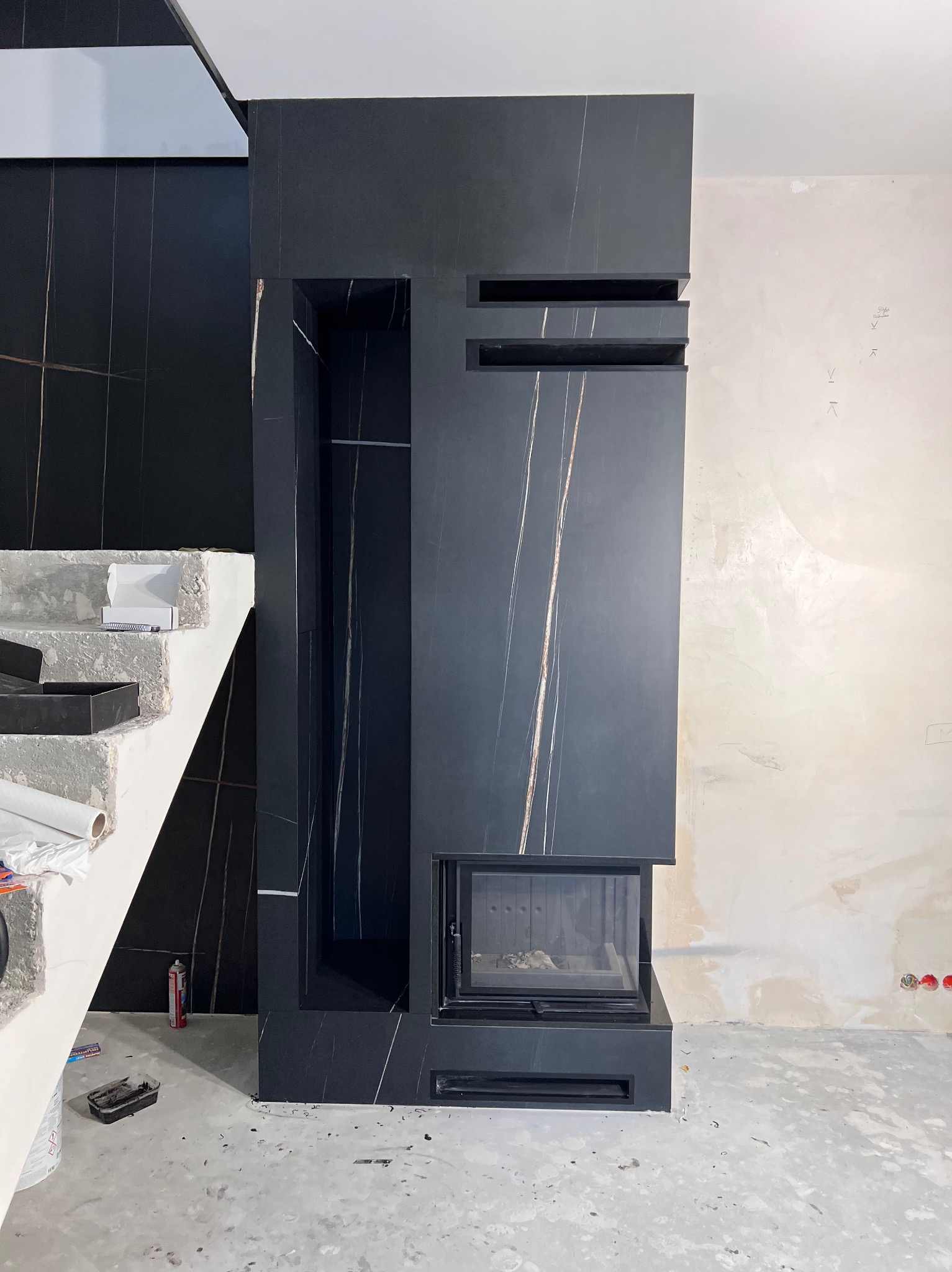 Nowoczesny kominek obudowany czarnym marmurem z wyraźnym użyleniem, w trakcie montażu, widoczne niedokończone ściany i schody z narzędziami.