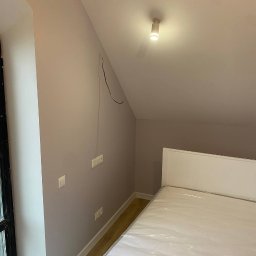 Komfort Bud - Pokój na poddaszu po malowaniu ścian na jasny fiolet, widoczne gniazdka elektryczne, wystające kable, białe łóżko z folią ochronną i drewniana podłoga.