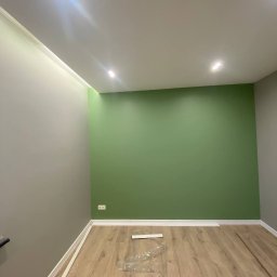 Komfort Bud - Świeżo pomalowane wnętrze z zieloną ścianą i panelami podłogowymi w trakcie układania, widoczne listwy przypodłogowe i folia ochronna na podłodze.