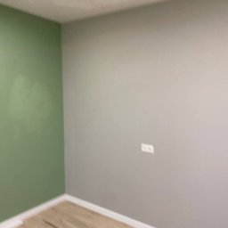 Komfort Bud - Pomieszczenie po malowaniu ścian na dwa kolory: zielony i szary, z ułożonymi panelami podłogowymi i zamontowanym gniazdkiem elektrycznym.