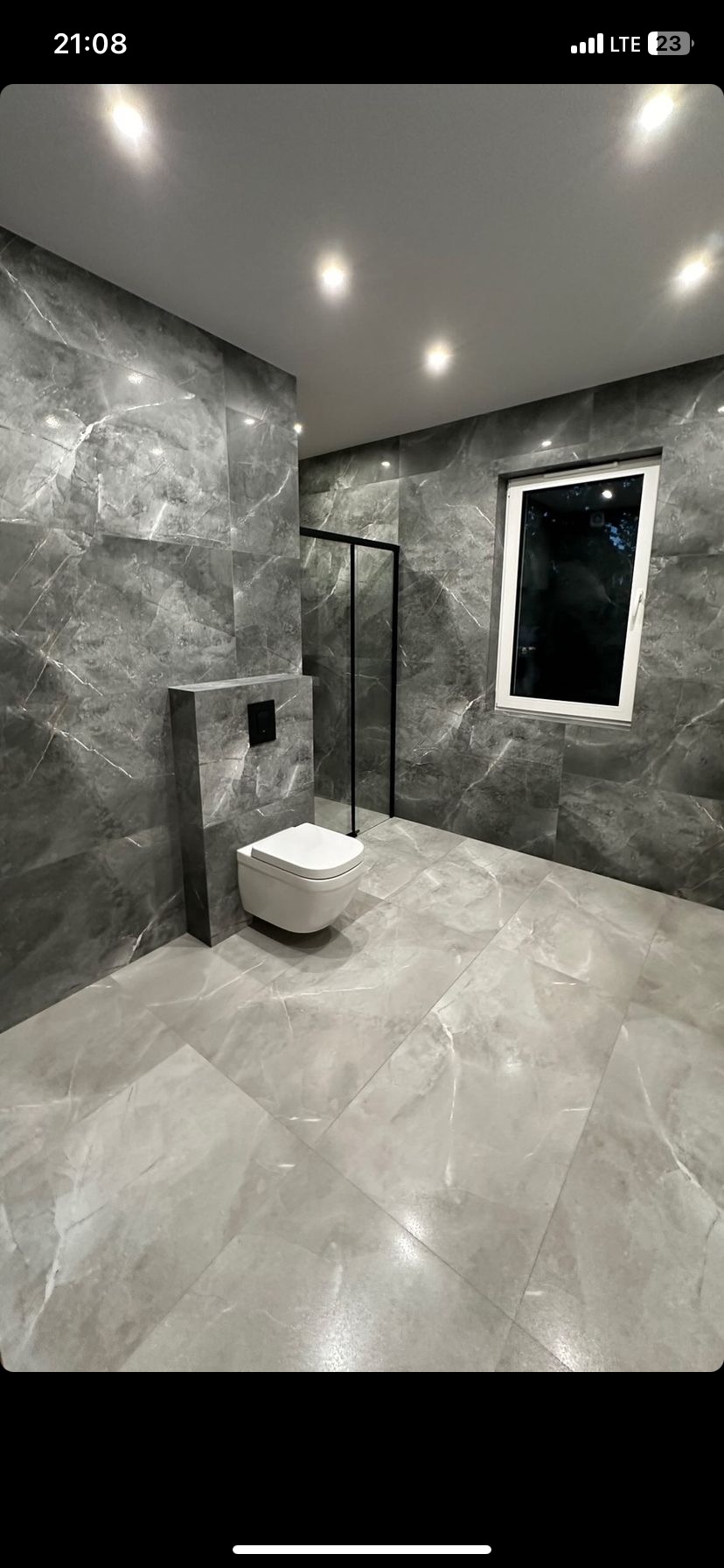 Nowoczesna łazienka z szarymi płytkami imitującymi marmur, podwieszana toaleta z czarnym przyciskiem i czarna kabina prysznicowa.