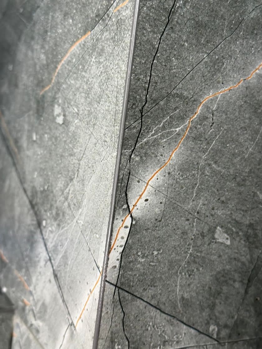 Szara płytka imitująca kamień z wyraźnymi czarnymi i złotymi żyłkami, ułożona z wąską, srebrną fugą, widziana pod kątem.