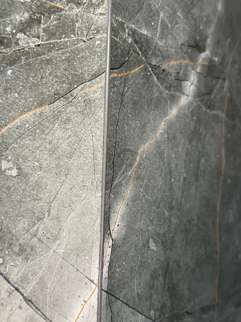 Szczegółowe ujęcie szaro-zielonych płytek imitujących marmur z wyraźnym użyleniem w odcieniach pomarańczy i bieli, ułożonych z wąską fugą.