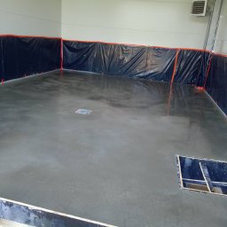 EsteRox Posadzki Przemysłowe - Świeżo wylana, gładka posadzka betonowa w hali, zabezpieczona folią przy ścianach. Widoczny odpływ liniowy i fragment szalunku. Butelka z wodą w rogu.