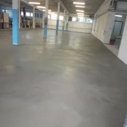 Gładka, szara posadzka przemysłowa w hali z niebieskimi słupami i rzędami lamp. Widoczne okna w tle i fragmenty wyposażenia. Świeża, r&oacute;wna powierzchnia.