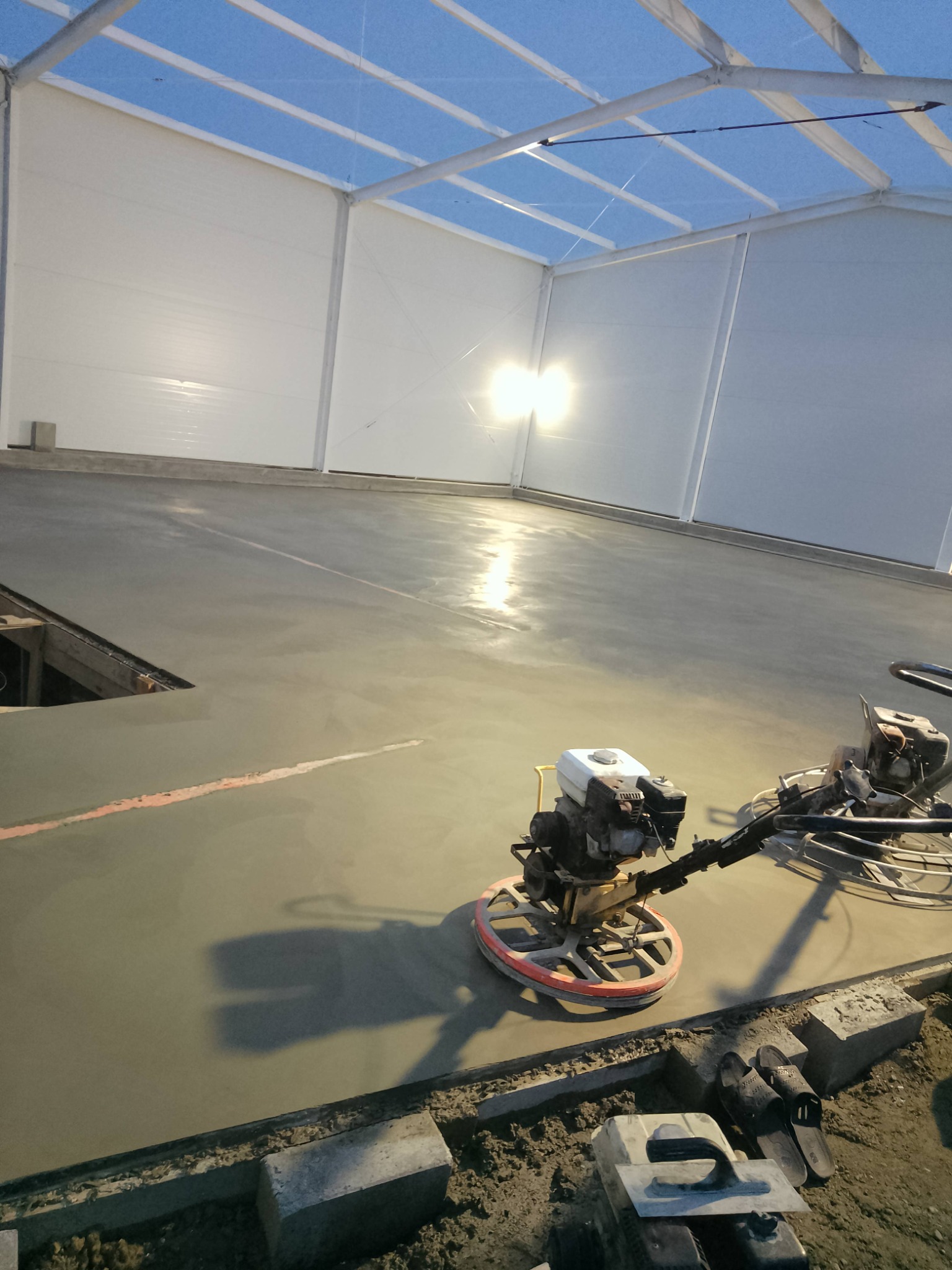 Zacieraczka mechaniczna w hali z posadzką betonową w trakcie prac. Świeży beton z czerwoną linią, białe ściany hali i jasne oświetlenie.