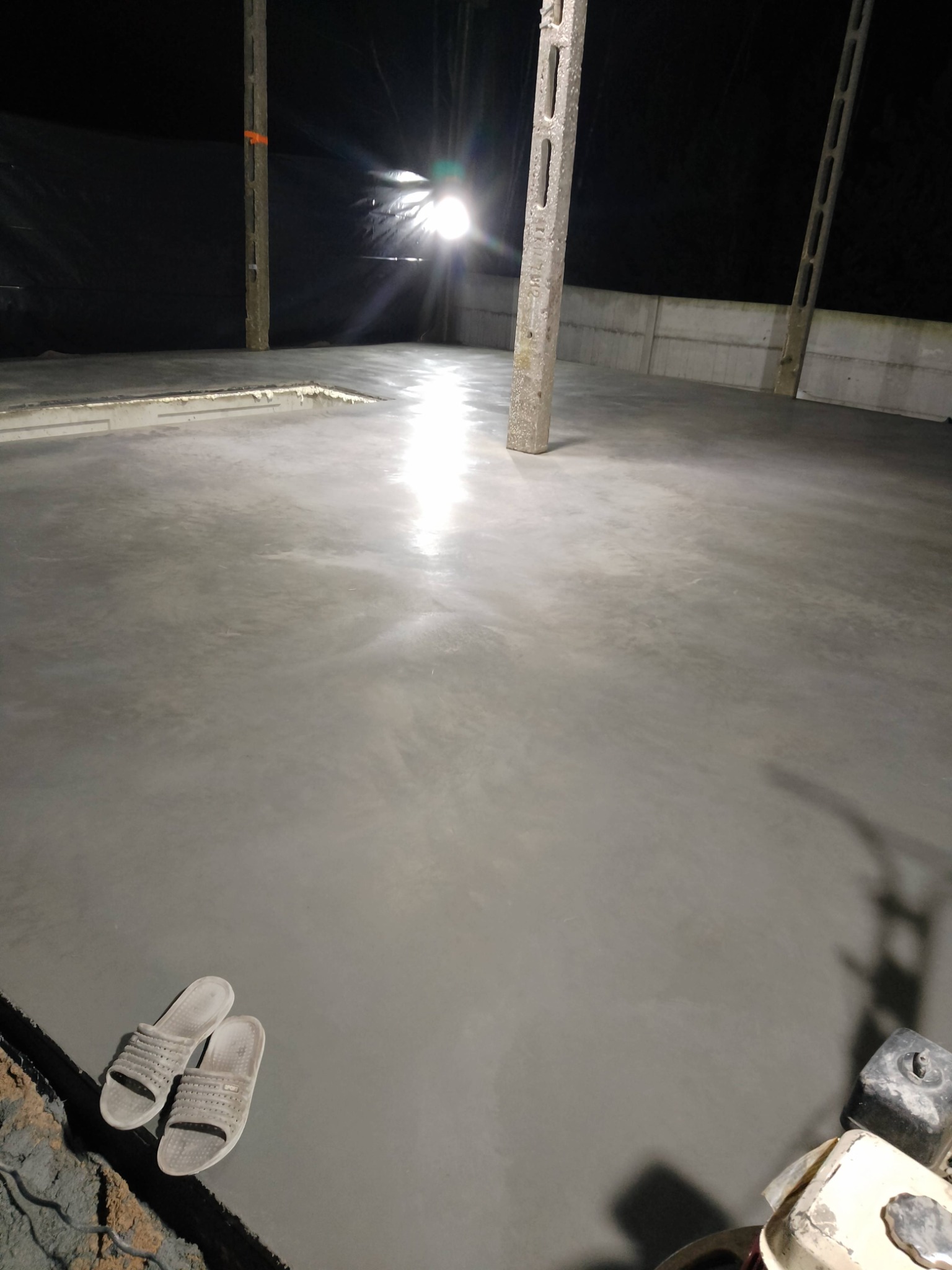Świeżo wylana, gładka posadzka betonowa w hali, z widocznymi słupami konstrukcyjnymi i odbiciem światła. W rogu leżą klapki i fragment maszyny.
