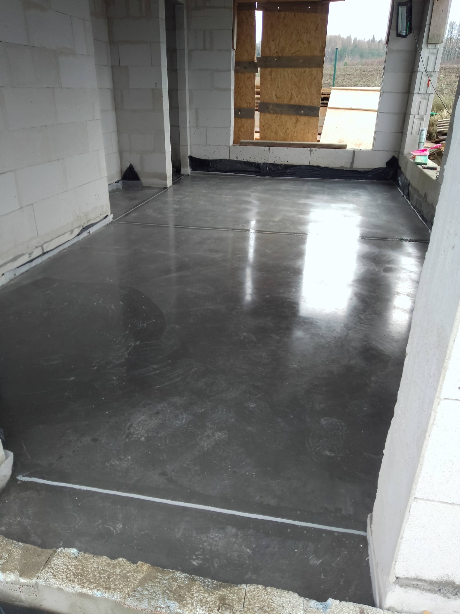 Wylana, gładka posadzka w surowym wnętrzu budynku z białych bloczków. Widoczne szczeliny dylatacyjne i folia wzdłuż ścian. Odbicia światła na mokrej powierzchni.