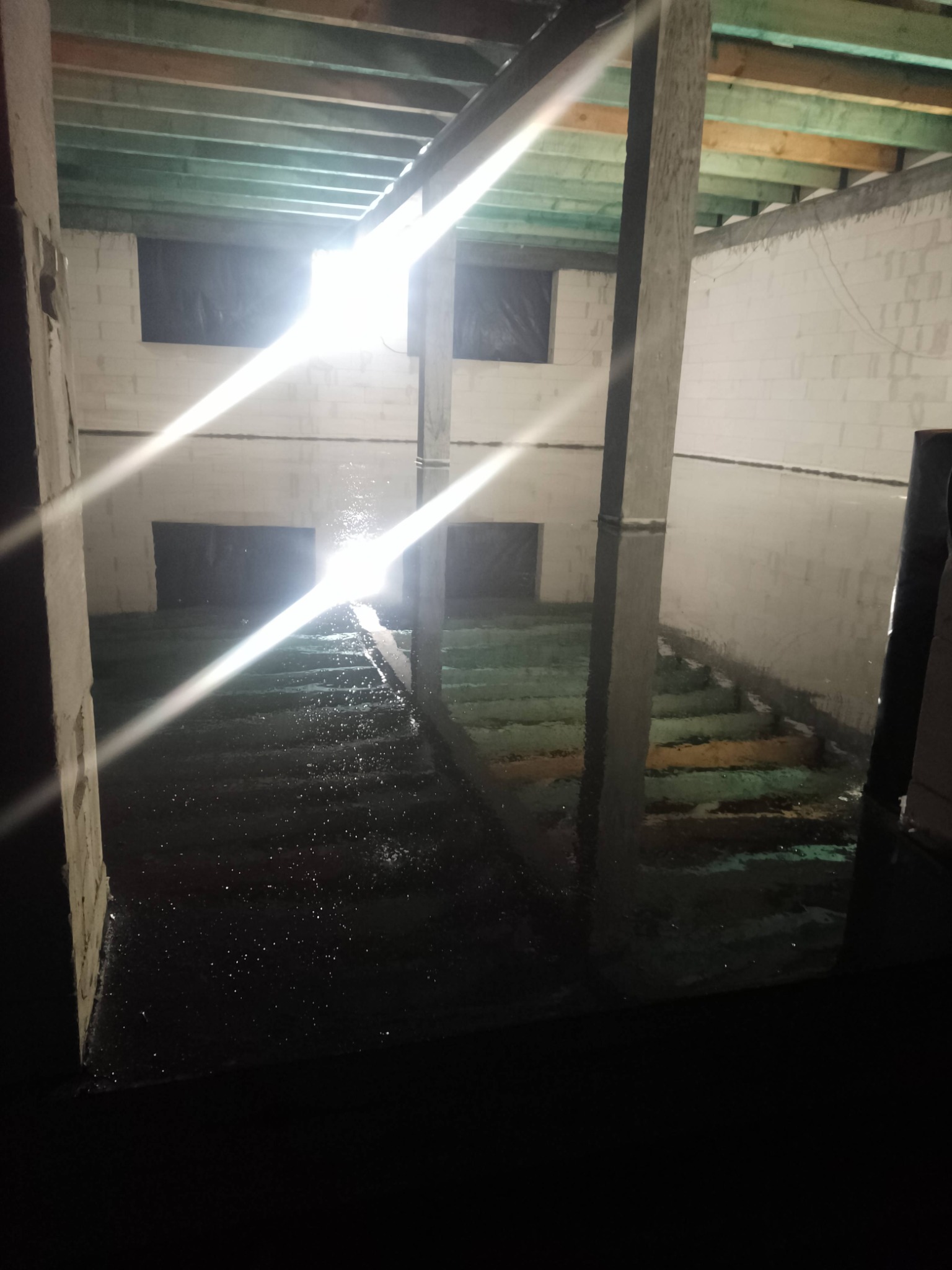 Wnętrze w trakcie budowy z zalaną posadzką, widoczne ściany z bloczków i drewniany strop. Mocne światło wpadające przez okna tworzy refleksy na wodzie.