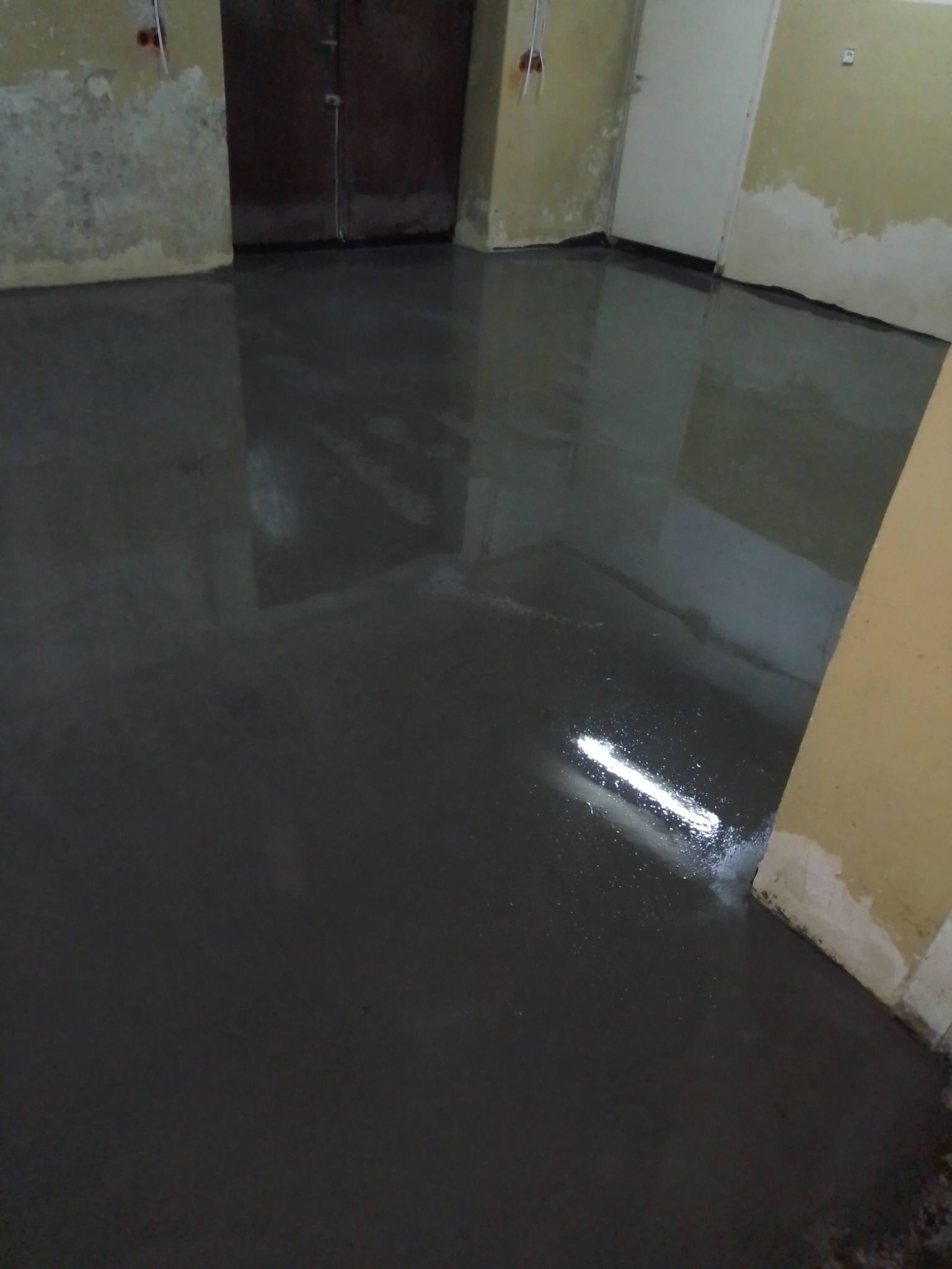 Świeżo wylana, ciemna posadzka z połyskiem w pomieszczeniu z surowymi ścianami. Widoczne ślady po wcześniejszych pracach budowlanych.
