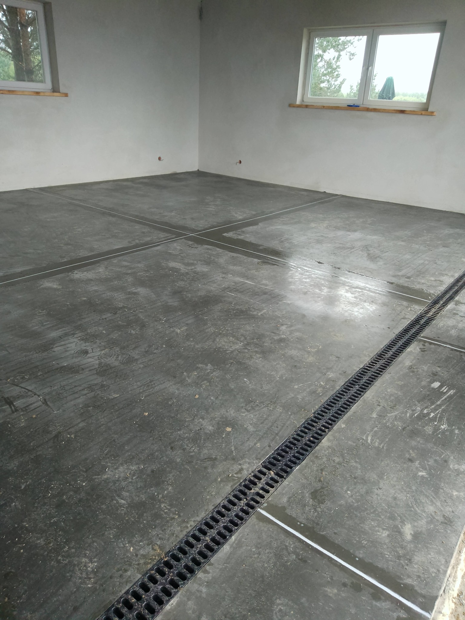 Szara, wilgotna posadzka przemysłowa z drenażem liniowym w nowym budynku. Widoczne fugi dylatacyjne i naturalne światło z okien.